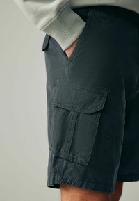 Dunkelgraue Cargo-Shorts aus einem Baumwoll-Mischgewebe, mit Seitentaschen und einer strukturierten Oberfläche. Der Bund ist mit einem Knopfverschluss verstellbar.