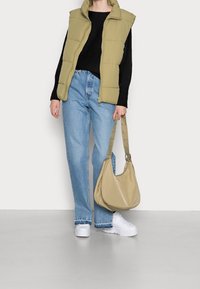 Khaki Steppweste über einem schwarzen Pullover, kombiniert mit hellblauen weit geschnittenen Jeans, weißen klobigen Sneakers und einer beigefarbenen Schultertasche mit Nieten.