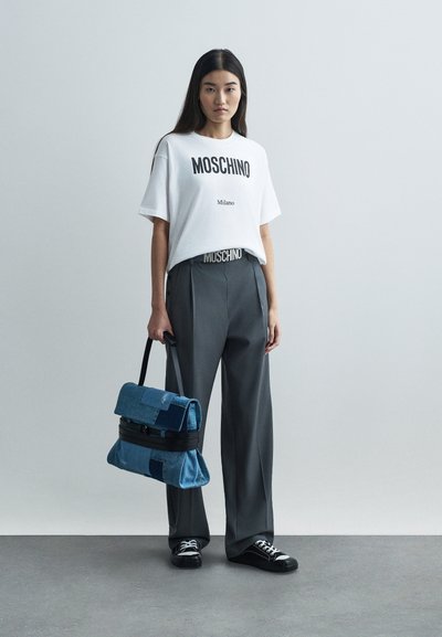 Woman wearing white Moschino T-shirt, gray wide-leg pants with Moschino belt, holding blue patchwork handbag, and black sneakers on gray floor.