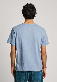 Šviesiai mėlyna medvilninė marškinėlių su apvalia iškirpte ir trumpomis rankovėmis, turinti laisvą siluetą ir šiek tiek nusidėvėjusias kraštines. Parodyta nugaros pusė.