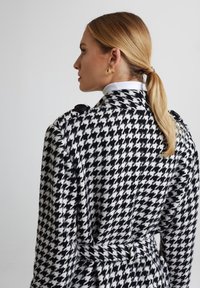 Czarny i biały płaszcz w wzór houndstooth z paskiem, dużymi guzikami na ramionach i kołnierzowym dekoltem. Miękki, teksturowany materiał.