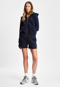 Navy zip-up hettegenser med frontlommer, kombinert med matchende shorts. Hettegenseren har en logo; modellen har på grå joggesko og hvite ankelsokker.