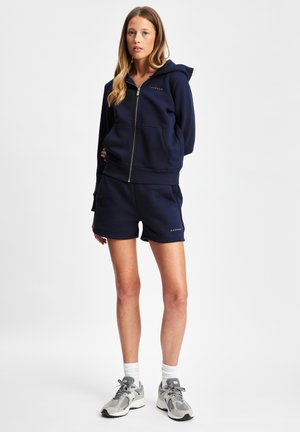 Navy zip-up hettegenser med frontlommer, kombinert med matchende shorts. Hettegenseren har en logo; modellen har på grå joggesko og hvite ankelsokker.