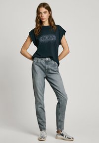 Jūrinis marškinėliai su trumpomis suktomis rankovėmis ir sidabrinėmis grandinėlėmis užrašyje "PEPE JEANS", derinami su šviesiai pilkais džinsais ir metaliniais sportbačiais.