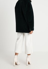 Musta keinokarvainen takki, jossa korkea kaulus, yhdistettynä valkoisiin leveälahkeisiin lyhyisiin housuihin ja vaaleisiin nilkkureihin. Sileä pinta näkyy.