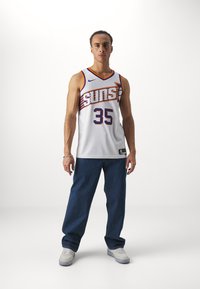 Maillot de basket-ball blanc avec des accents orange et violet, numéro 35, portant un pantalon en denim et des baskets blanches, le tout sur un fond uni.