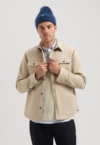 Beige uldblandet skjortejakke med to brystlommer og knaplukninger, lagdelt over en tekstureret strikket sweater og en stribet skjorte. Blå beanie-cap.