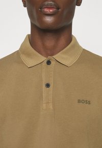 Tan polo skjorta med klassisk krage, två svarta knappar och diskret logotyp. Tyget har en strukturerad yta.