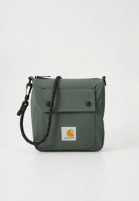 Mala verde de nylon para usar ao ombro com aba frontal, dois botões de pressão e alça preta. Apresenta um emblema com design em laranja e branco.