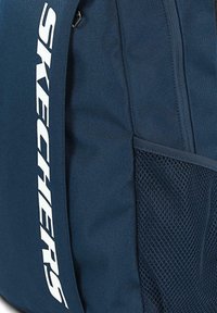 Mochila azul marina con tela texturizada, que cuenta con un bolsillo de malla lateral y un gran logo blanco de "SKECHERS" en la correa.