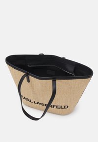 Taška typu tote z béžového tkaného materiálu s černými koženými popruhy a doplňky, na jedné straně černé logo a uvnitř kapsa na zip.