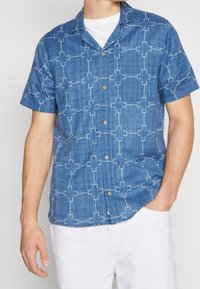 Modrá džínová košile s krátkými rukávy a zapínáním na knoflíky, zdobená bílým geometrickým vzorem a hnědými knoflíky, s volným střihem.