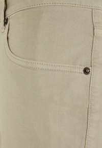 Una tela de mezclilla beige forma un bolsillo ajustado con un ligero brillo. Presenta un botón metálico decorativo y costuras visibles a lo largo de la costura.
