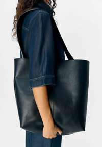 Une personne portant un jean bleu foncé tient un grand sac cabas en cuir noir avec une bandoulière contre un fond blanc.