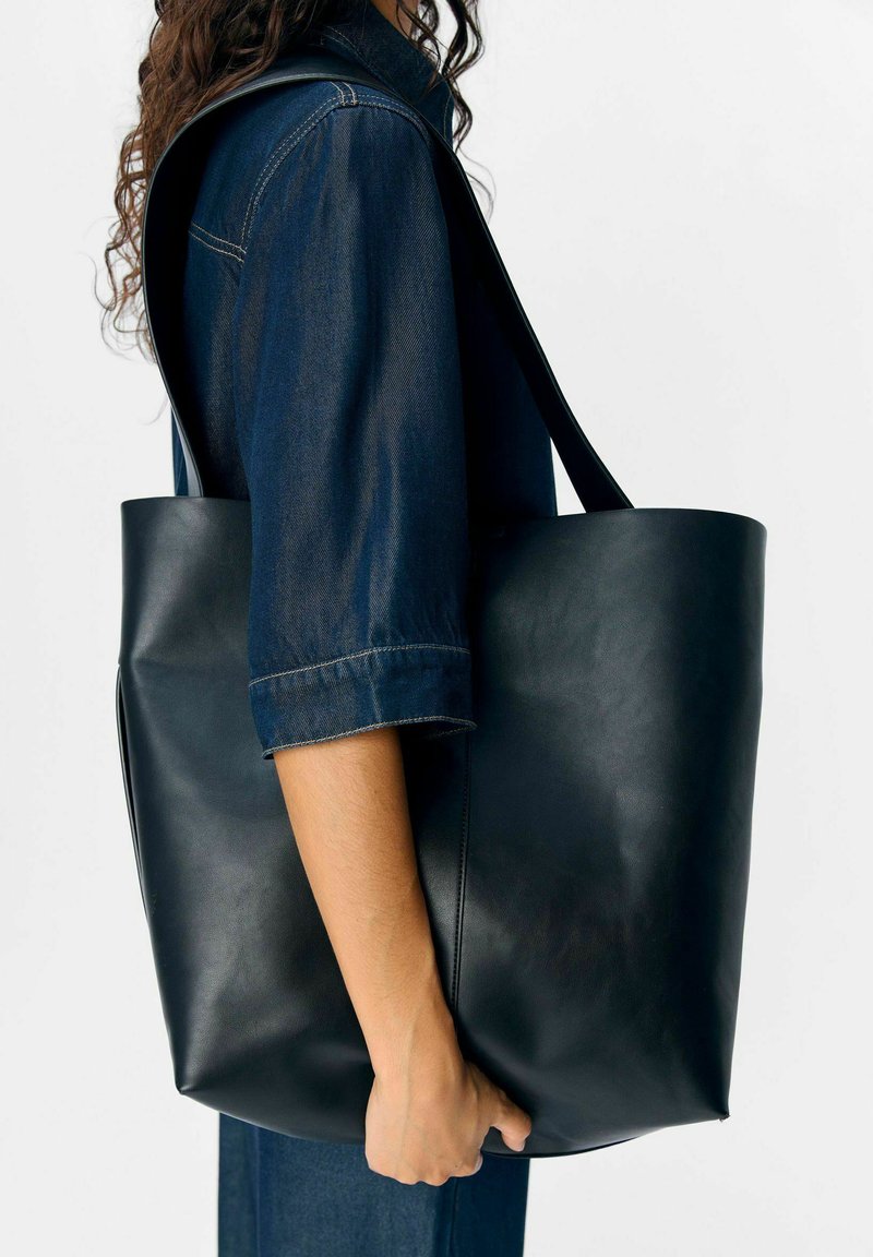 Une personne portant un jean bleu foncé tient un grand sac cabas en cuir noir avec une bandoulière contre un fond blanc.