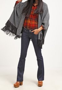 Graues Strickponcho mit fransigen Kanten, getragen über einem rot-schwarzen karierten Hemd, kombiniert mit dunklen, ausgestellten Jeans und beigefarbenen Plateauschuhen.
