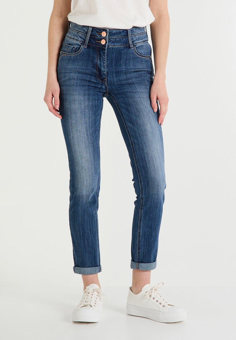 Jeans de mezclilla azul oscuro de cintura alta, con dos bolsillos frontales, puños enrollados y acentos de botones de bronce. Combinados con zapatillas blancas.