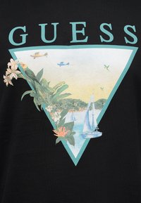 Το λογότυπο GUESS με τροπικά φυτά, ένα παπαγάλο, ιστιοφόρα σε νερό, μακρινά λόφους, μικρά αεροπλάνα και κολίβρια μέσα σε τρίγωνο με πράσινο περίγραμμα.