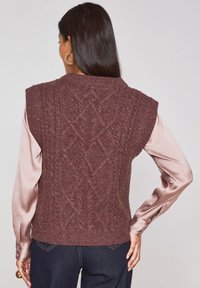 Maglione senza maniche in maglia borgogna con motivo a treccia testurizzato. Abbinato a una blusa satinata chiara a maniche lunghe, indossata sopra jeans di denim scuri.