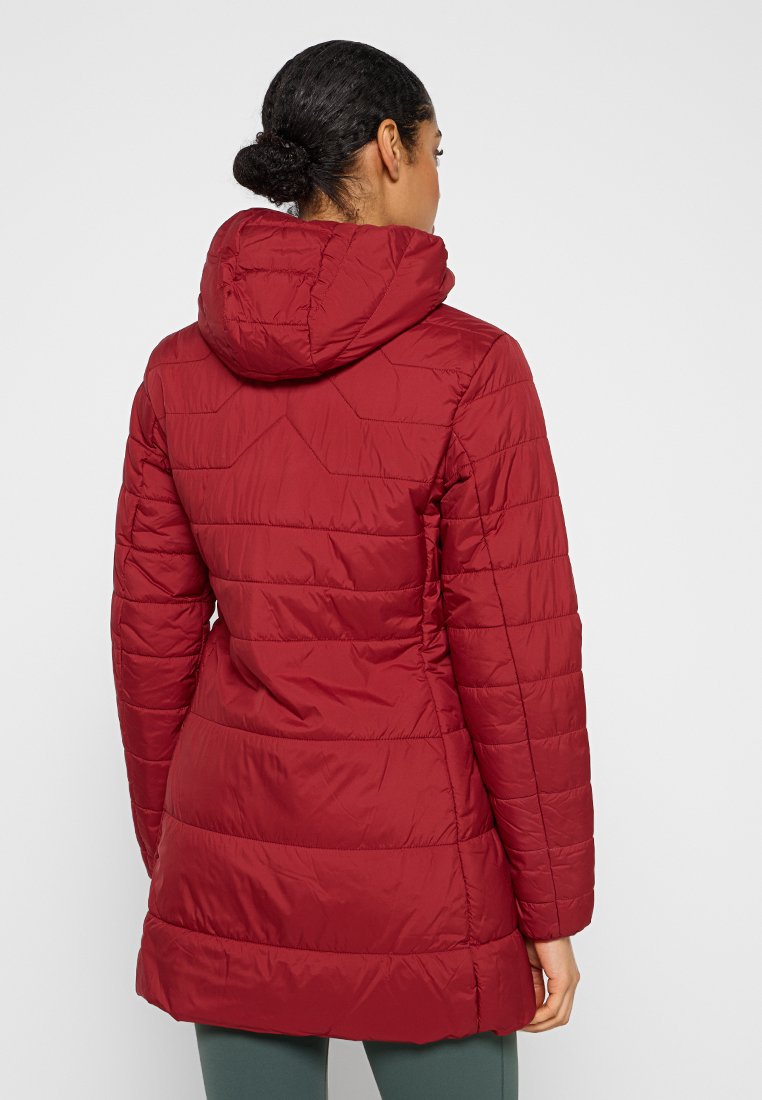 Frau mit dunklem Haar, das zu einem Dutt gebunden ist, trägt eine lange, gesteppte rote Kapuzenjacke und steht mit dem Rücken zu einem schlichten, hellen Hintergrund.