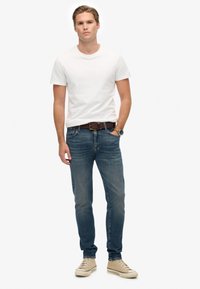 Joven de pie contra un fondo blanco, vistiendo una camiseta blanca, jeans azules, cinturón marrón, zapatillas beiges y un reloj de pulsera.
