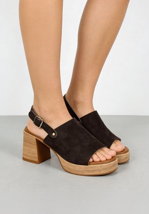 Pies con sandalias de plataforma abiertas en la punta de ante negro con talones de bloque de madera sobre una superficie gris.