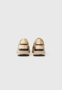 Des sneakers beiges avec des bords noirs et des semelles épaisses, vus de l'arrière sur un fond neutre.