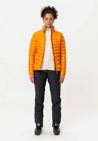 Oranje geïsoleerd jack met doorgestikte textuur over een lichtgekleurde top met rits, gecombineerd met zwarte broek en skischoenen, rechtopstaand.