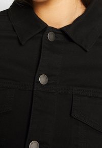 Černá džínsová bunda s límcem, kovovými cvoky, náprsními kapsami a viditelnými švy. Hladká textura a přiléhavý střih.