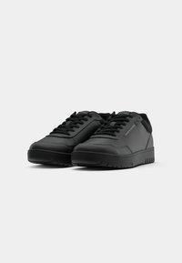 Paire de baskets basses noires avec lacets et marquage subtil sur les côtés et la languette, sur un fond gris clair uni.