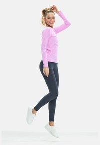 Rosa langärmliges Sportoberteil, kombiniert mit dunklen Leggings und weißen Turnschuhen. Das Outfit ist eng geschnitten und betont ein sportliches Design.