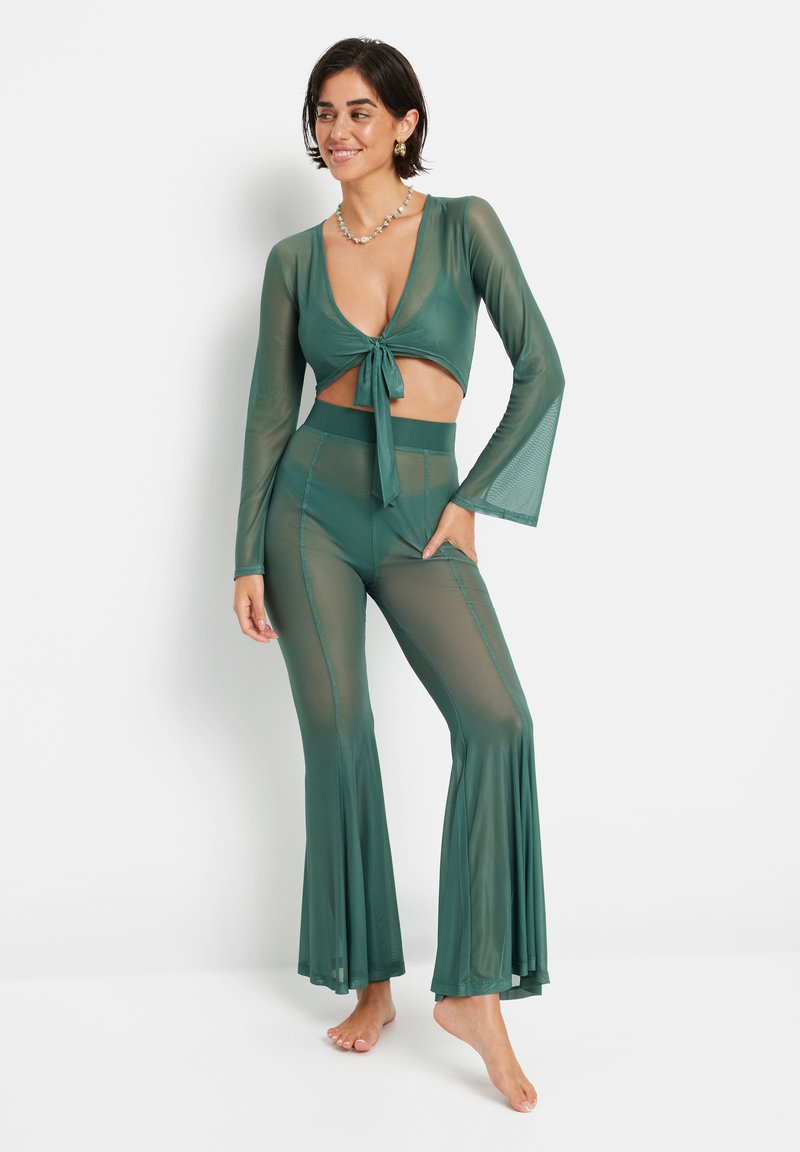 Completo in rete a due pezzi di colore verde scuro; crop top a maniche lunghe con nodo frontale e pantaloni svasati con una texture liscia e orli larghi.