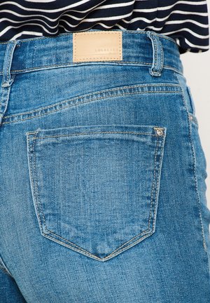 Jeans in denim blu con un'etichetta in pelle, che mostrano il dettaglio della tasca posteriore e le cuciture. Il tessuto ha una finitura leggermente strutturata.