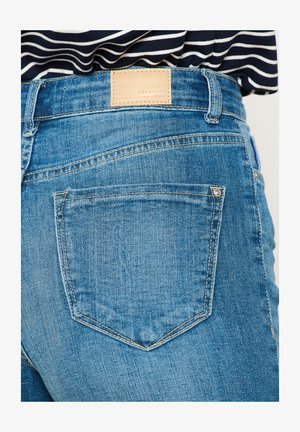 Jeans in denim blu con un'etichetta in pelle, che mostrano il dettaglio della tasca posteriore e le cuciture. Il tessuto ha una finitura leggermente strutturata.
