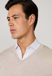 Mężczyzna z krótkimi ciemnymi włosami, ubrany w jasnobeżowy sweter z dekoltem w serek na białej koszuli z guzikami, patrzy w lewo na jednolitej tle.