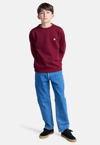 Boy stands with hands in pockets, wearing a burgundy sweatshirt, blue jeans, and black sneakers, set against a plain white background.