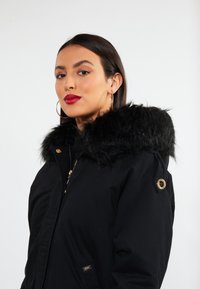 Sort parka-jakke med en falsk pelskrave, guldl�ås og hardware-detaljer. Har en lynlåse foran og en let glans på stoffet.