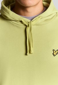 Ljusgul hoodie med en framfickad kangaroo-lomme, dragsko i huvan och en liten broderad logotyp i svart på vänster bröst. Material i bomull, slät textur.
