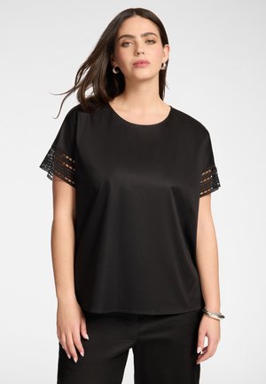 Kobieta ubrana w czarną bluzkę z krótkim rękawem z koronkowym zdobieniem przy rękawach i dopasowane czarne spodnie, stojąca na jednolitym tle.