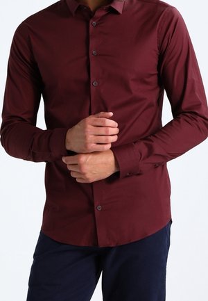 Man draagt een nauwsluitend bordeauxrood overhemd met lange mouwen en donkere blauwe broek, terwijl hij de mouwen aanpast met gekruiste handen voor zich.