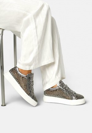 Piedi con sneakers argentate borchiate con suole bianche, abbinate a pantaloni bianchi larghi, appoggiati su una gamba di sedia metallica su sfondo bianco.