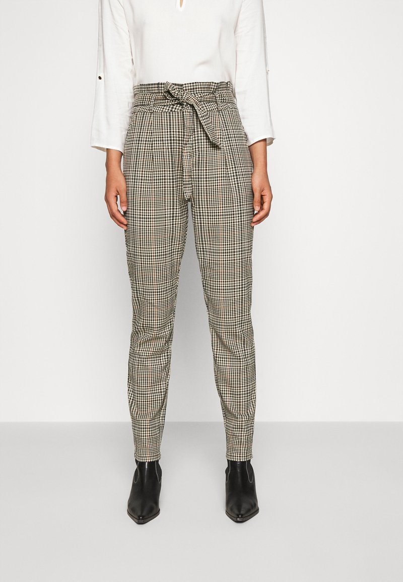 Femme portant un pantalon paperbag à carreaux taille haute avec ceinture nouée, un haut blanc à manches longues et des bottines noires.