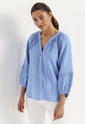 Vrouw met bruin haar in een staart, gekleed in een lichtblauw blouse met knopen en kantdetails, en een witte broek, tegen een effen witte achtergrond.