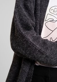 Tmavě šedý pletený kardigan s volným střihem a jemnou texturou, s širokým otevřeným předním dílem a žebrovanými manžetami, nošený přes bílou košili.