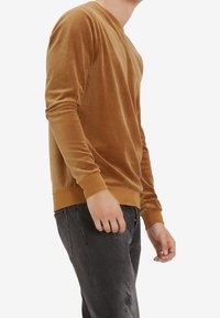 Sudadera larga de manga con cuello redondo en terciopelo marrón, con puños y dobladillo ajustados. Combinada con jeans oscuros desgastados. Textura suave.