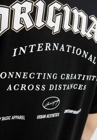 Sort t-shirt med hvid typografi, herunder "ORIGINAL" og andre tekstelementer. Glat tekstur med en tætsiddende pasform.