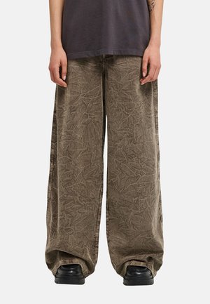 Persona che indossa pantaloni di velluto a coste marroni larghi che si raccolgono alle caviglie sopra scarpe nere, con le mani lungo i fianchi e una camicia scura.