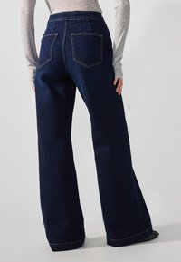 Tamno plave široke traperice s visokim strukom, s dva stražnja džepa i vidljivim šavovima, izrađene od denim tkanine.