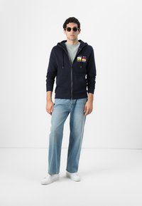 Sweat à capuche zippé marine avec des accents de logo, t-shirt gris, jeans bleu clair et baskets blanches. Modèle portant des lunettes de soleil noires, cheveux bouclés et coupés.