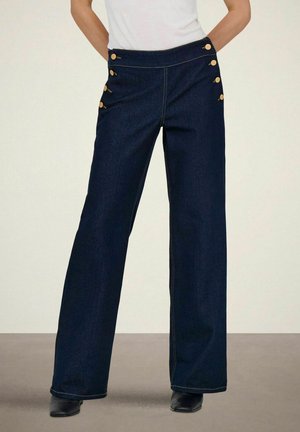 Nő magas derekú, sötétkék, széles szárú farmert visel arany gombokkal a csípő oldalán, és fekete bokacsizmát.